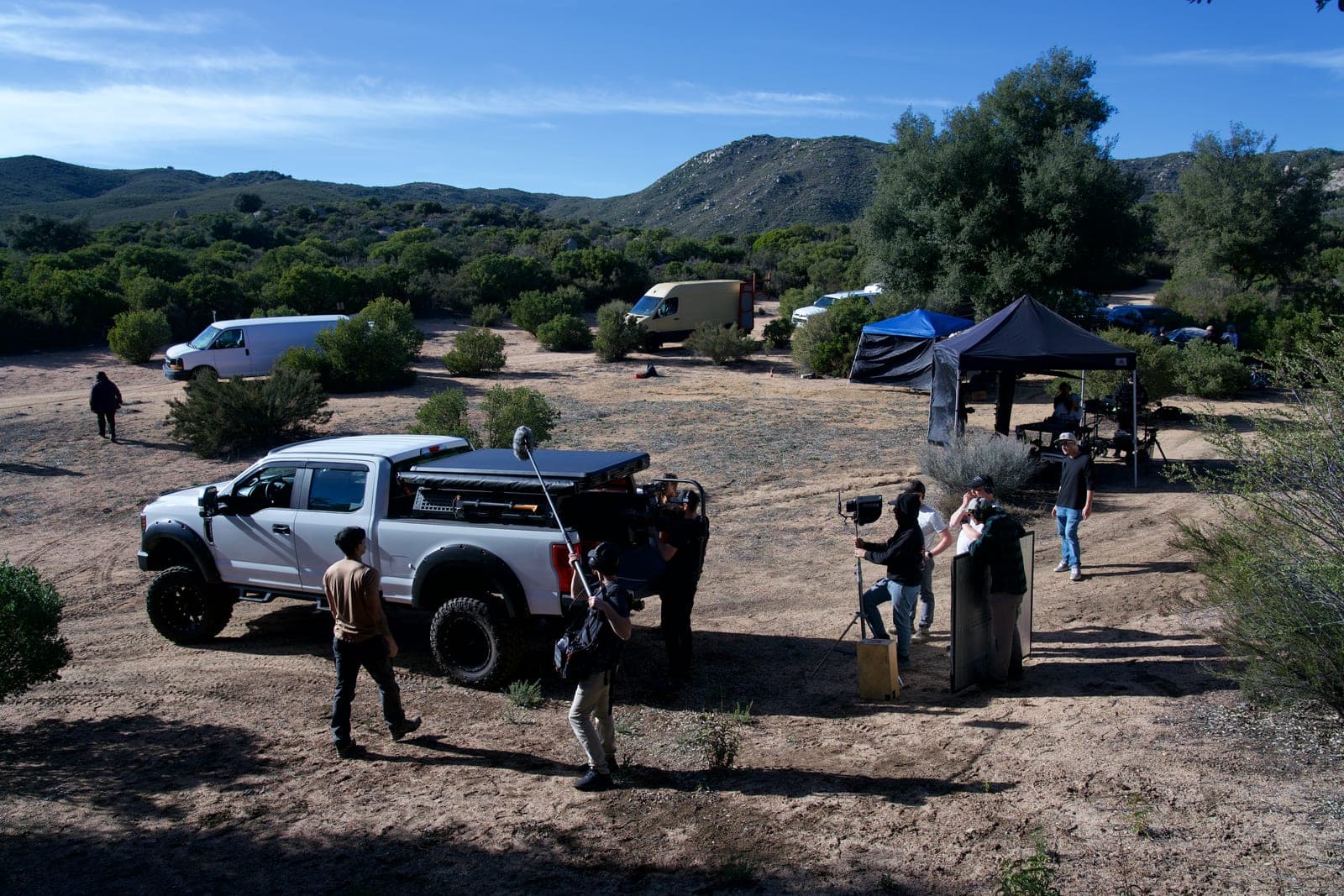 A wide of the full unit working in scrubland: truck, boom operator, video village under tents and vehicles at the treeline