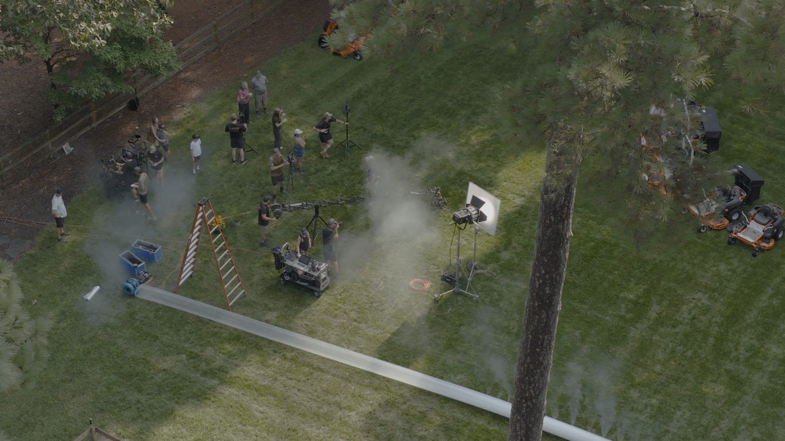 A drone plate looking down on a full unit working a lawn: crew, ladder, lighting stands, haze drifting across the grass and mowers staged at the treeline