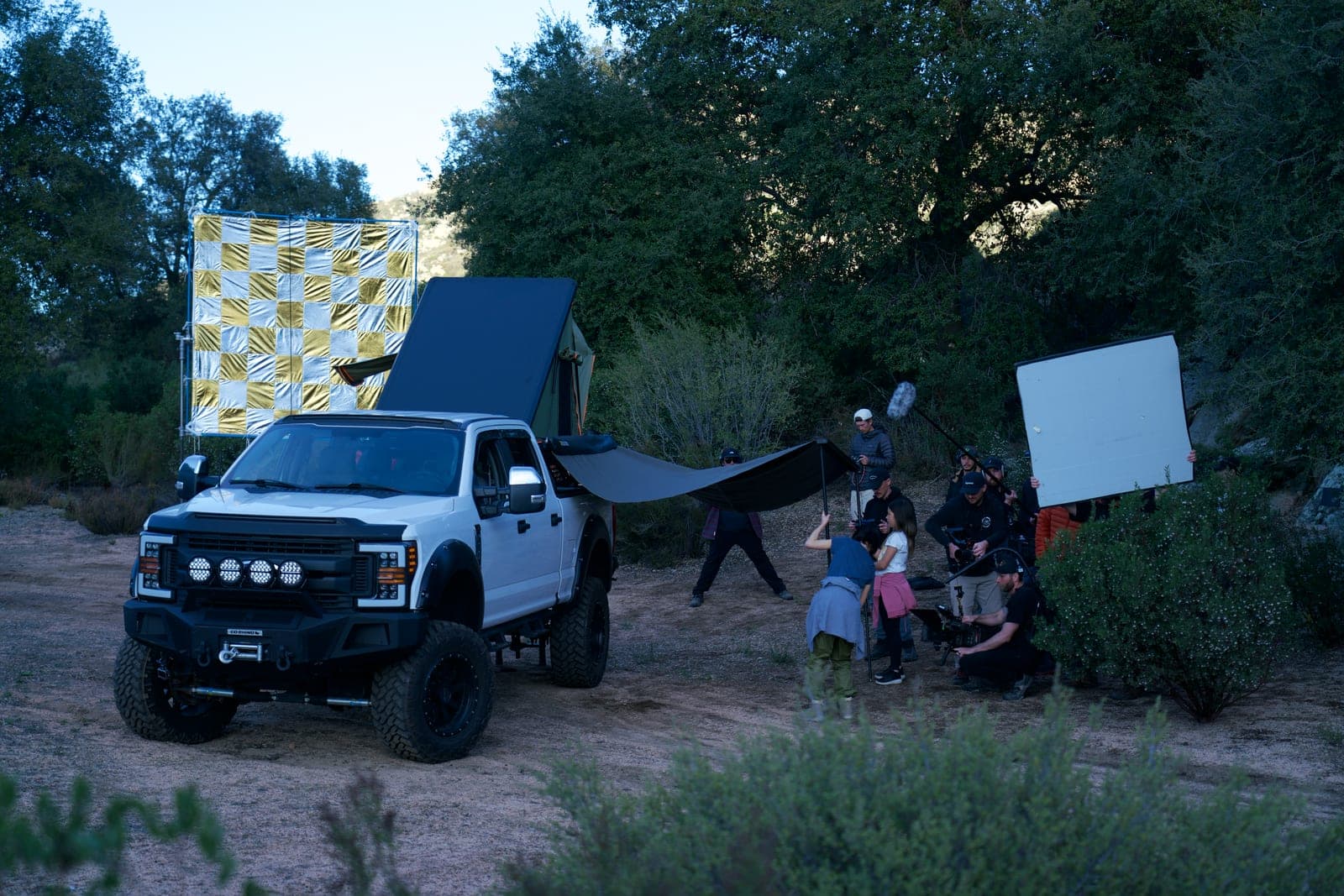 A pickup lit for a dusk shot in scrubland, crew working a bounce and a black flag either side of it