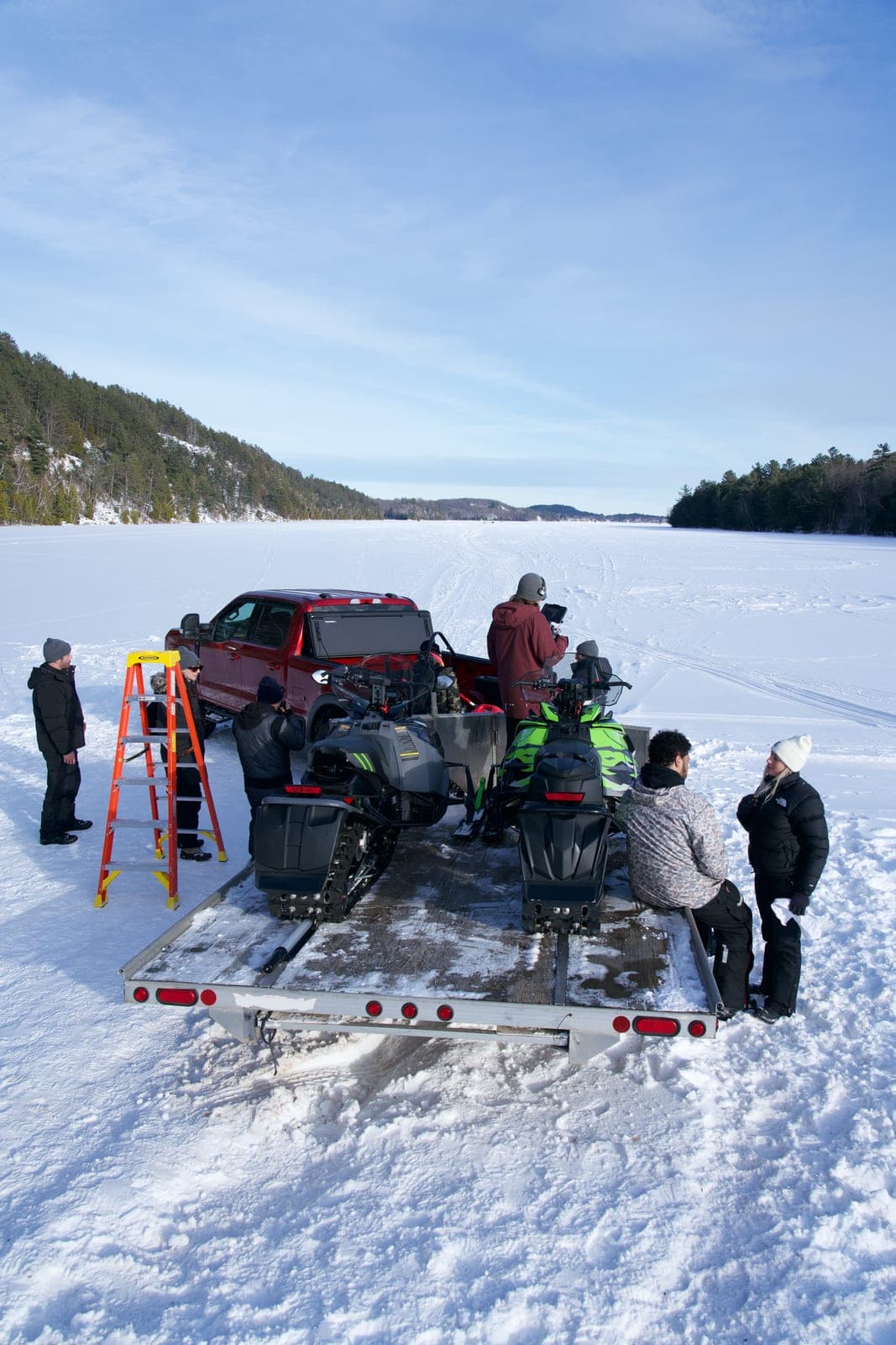 The unit loading snowmobiles off a trailer onto a frozen lake, truck and stepladder beside them