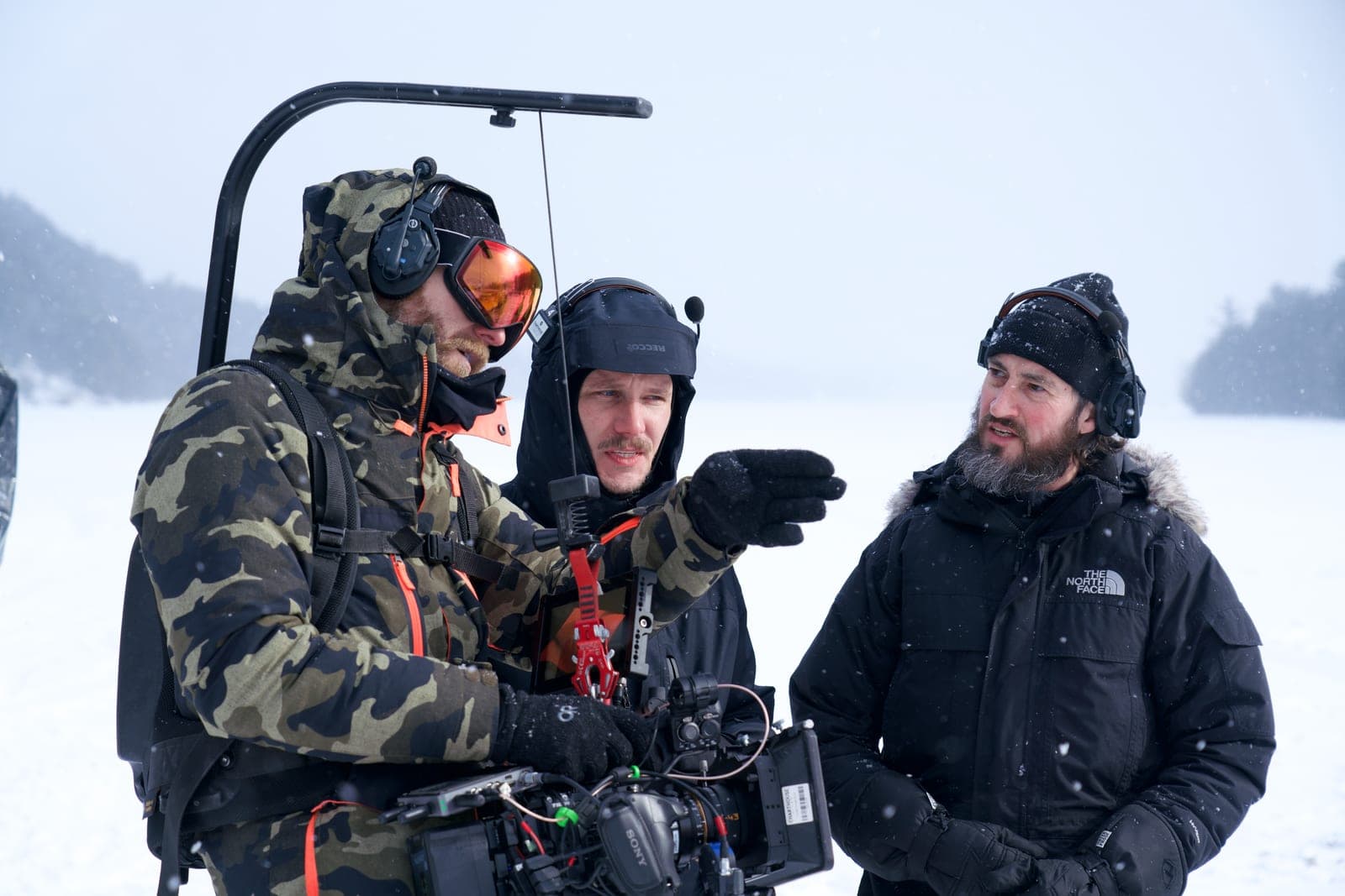Three crew in heavy winter gear talking through a shot on a frozen lake, camera rig on a gimbal between them