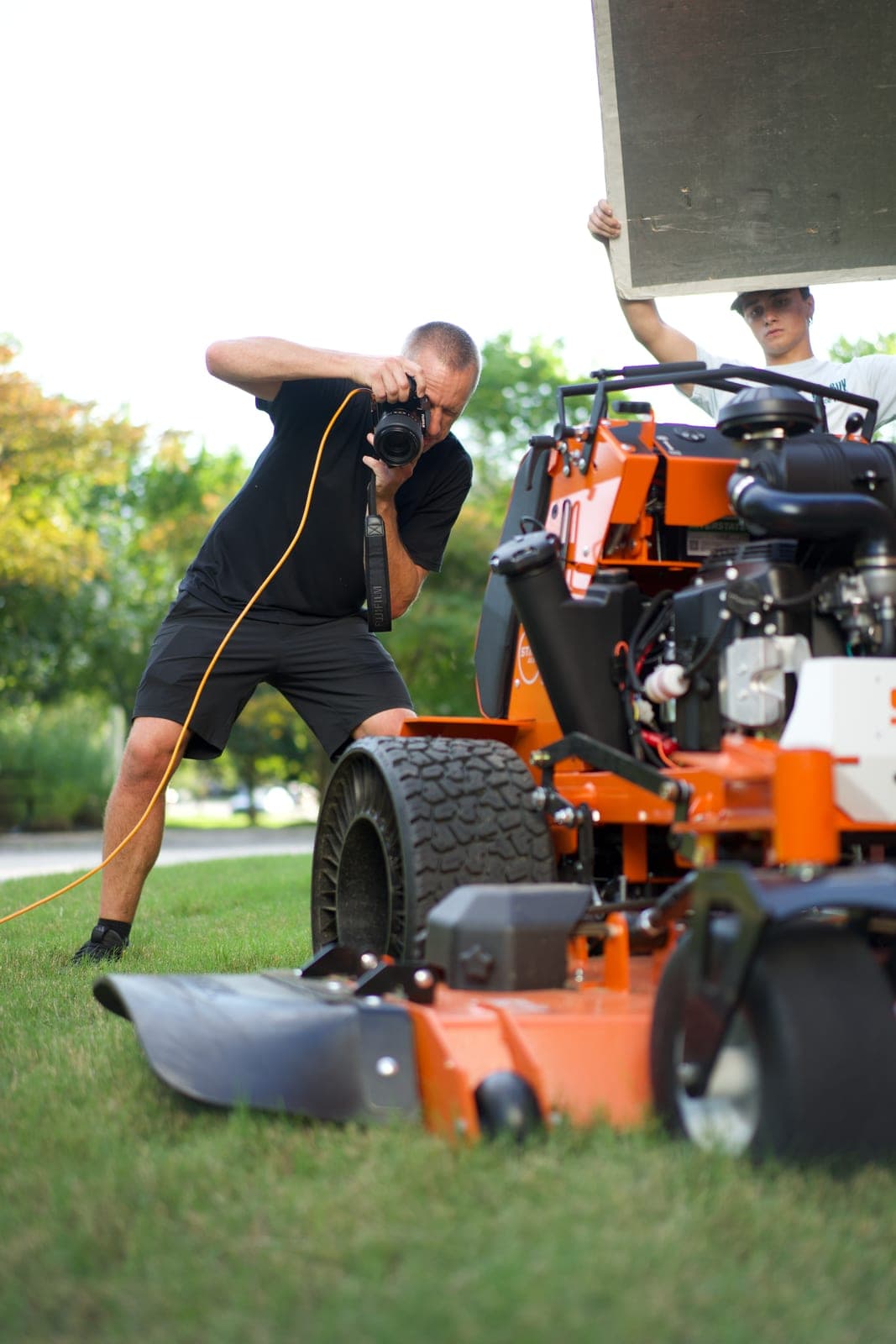 A stills photographer crouched low against a machine while a crew member holds a bounce board overhead
